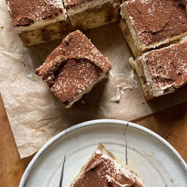 A slice of tiramisu tres leches cake on a plate with the full cake on the table