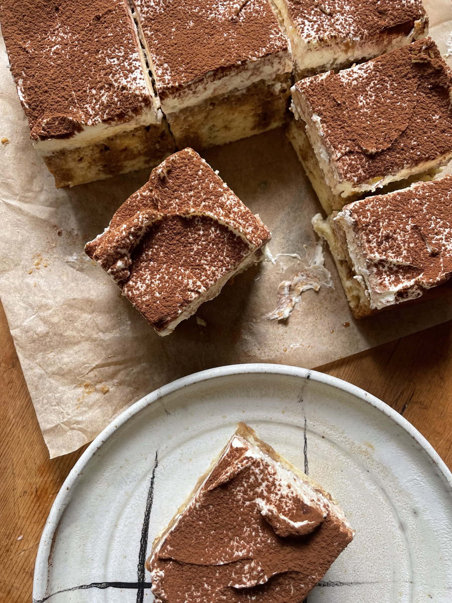 A slice of tiramisu tres leches cake on a plate with the full cake on the table