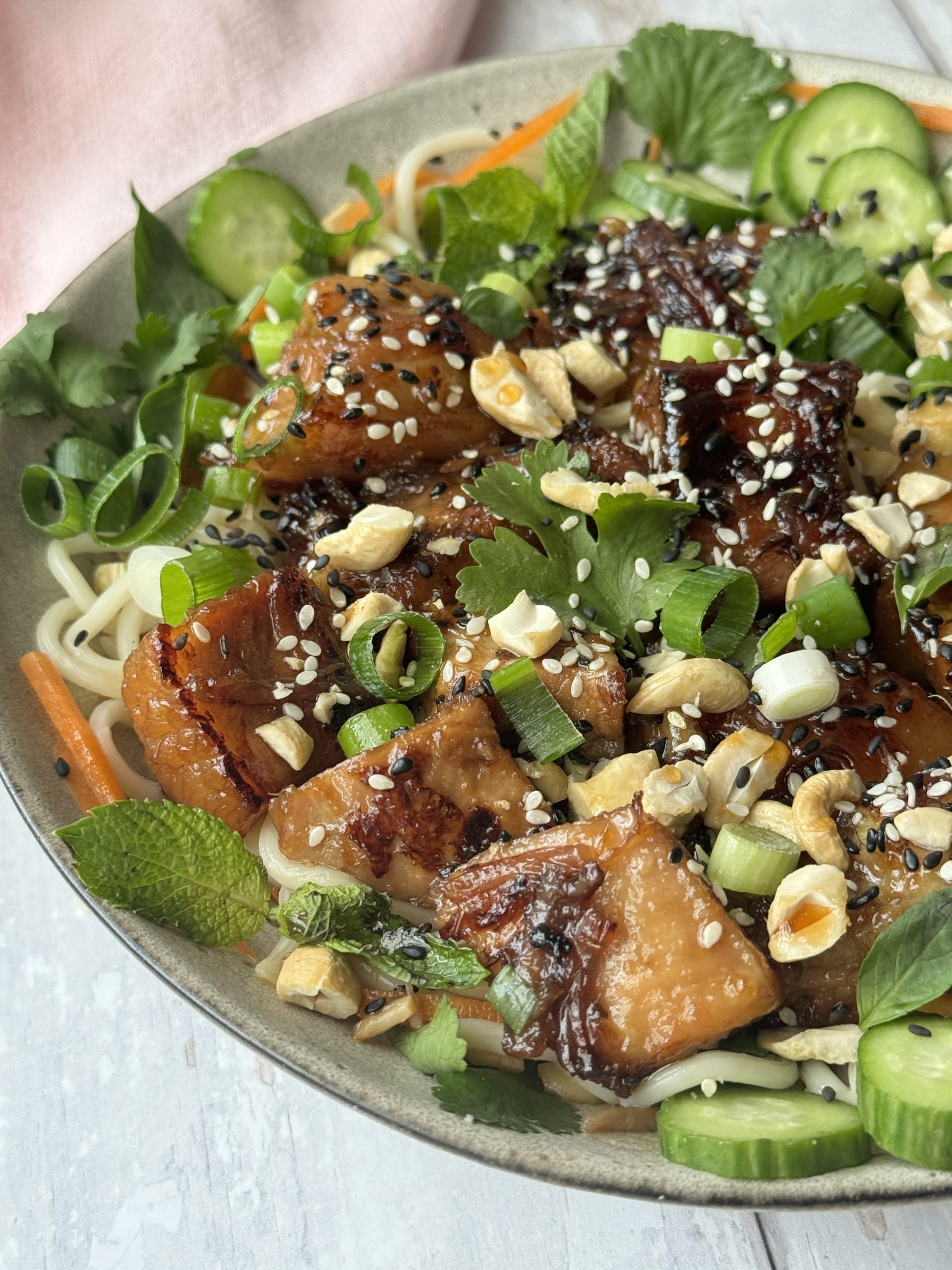 pork belly noodles in a white bowl decorated with herbs and cucumber