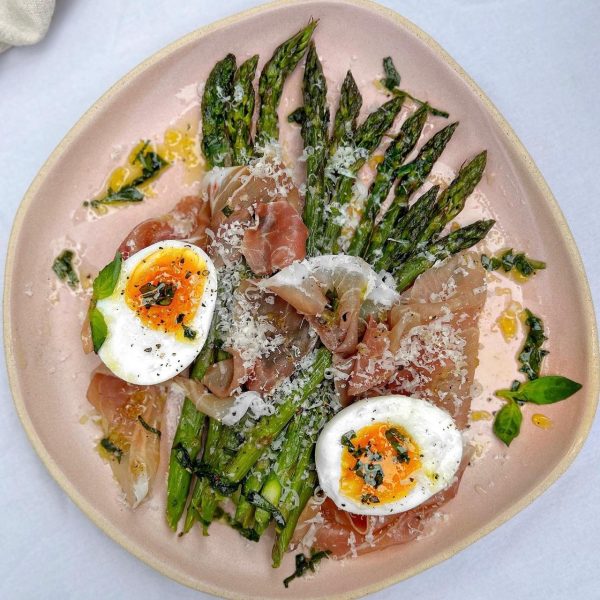 asparagus on a pink plate topped with parma ham, parmesan and a soft boiled egg