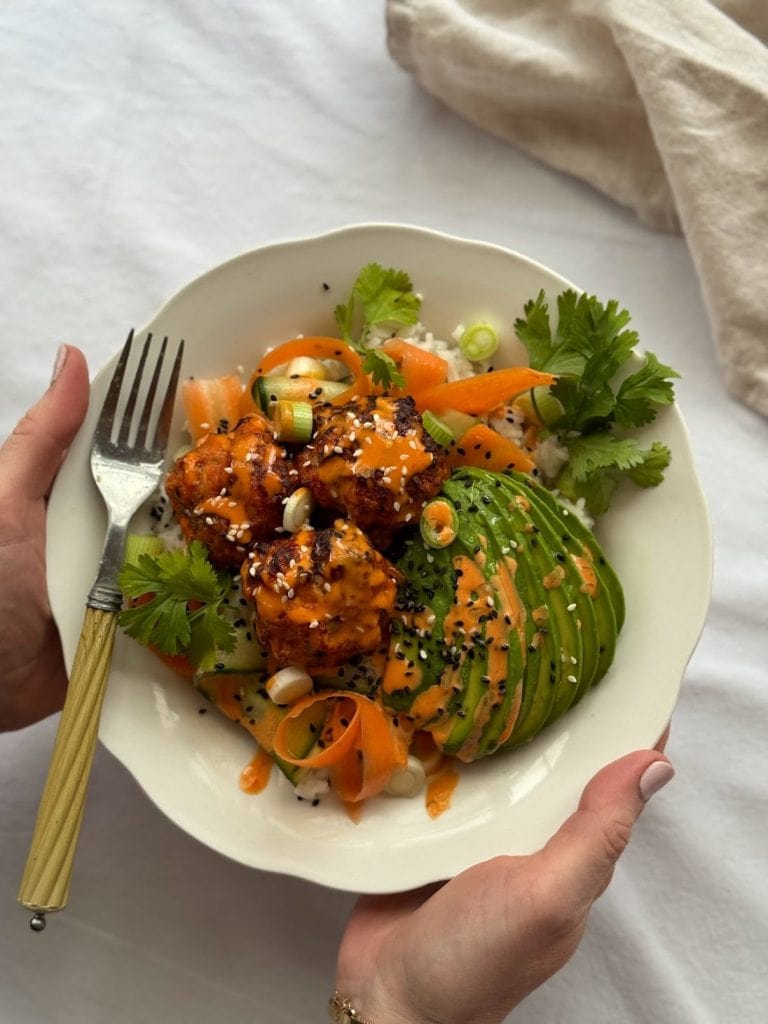 A woman holding a white bowl of spicy fishcakes served with gochujang mayo, avocado, carrot, cucumber, coriander and white rice.