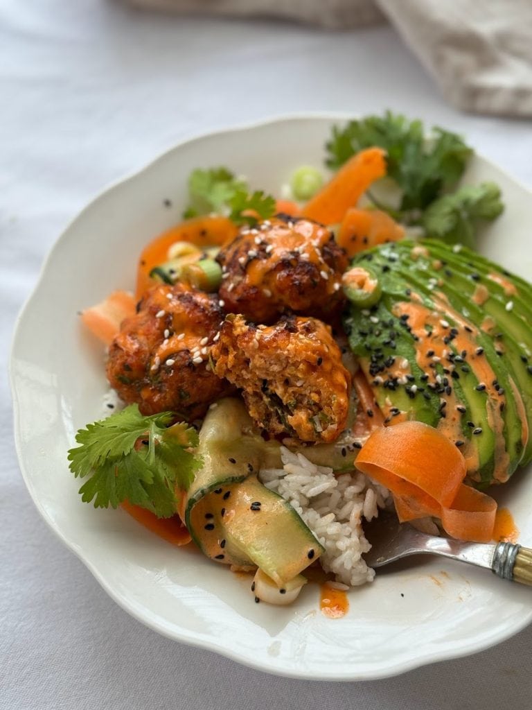 Close up of a bowl of salmon fishcakes served with cucumber ribbons and sliced avocado with a spicy mayo over rice. 