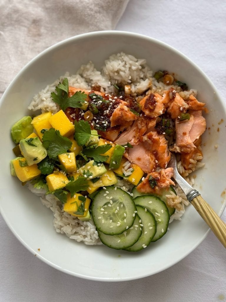 White bowl with a cucumber, teriyaki salmon and avocado mango salad served over white sushi rice.