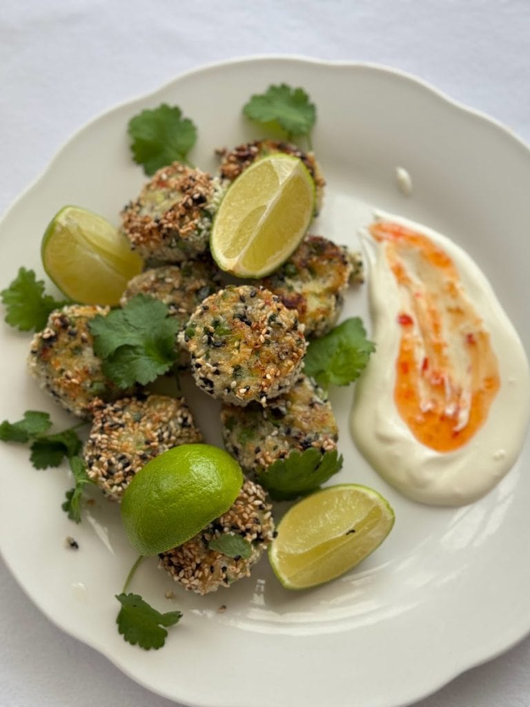 White plate of sesame seed coated fish cakes with lime wedges, fresh coriander and a creaky sweet chilli dip.