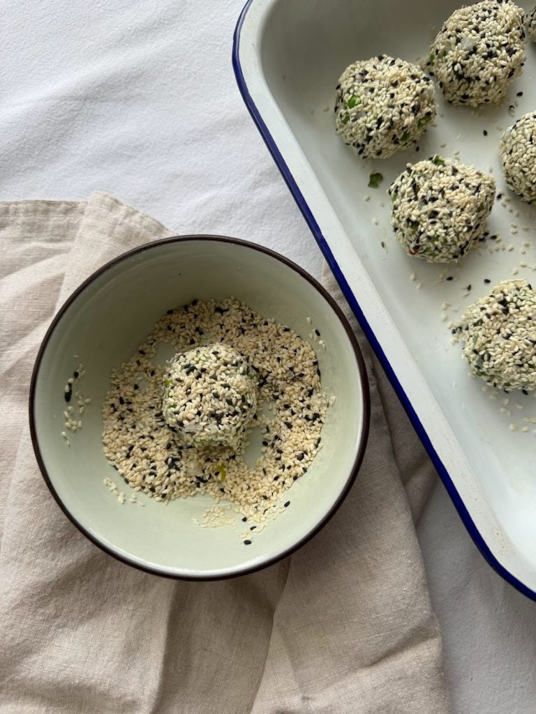 Fishcakes being rolled in a shallow bowl of mixed black and white sesame seeds. 