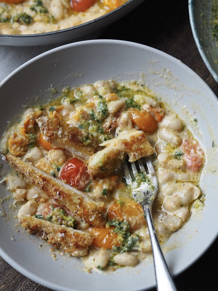 Sliced breaded chicken breast in a bowl of white beans with tomatoes and basil.