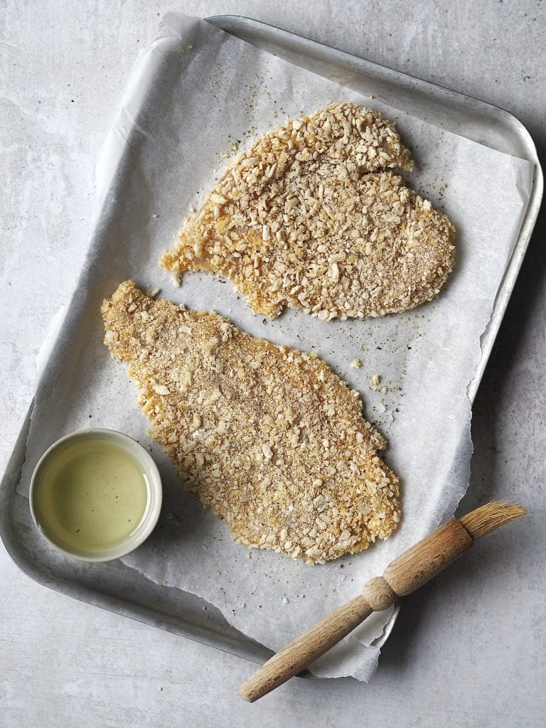 Breaded chicken cutlets on a lined baking tray  being brushed with a bowl of oil.