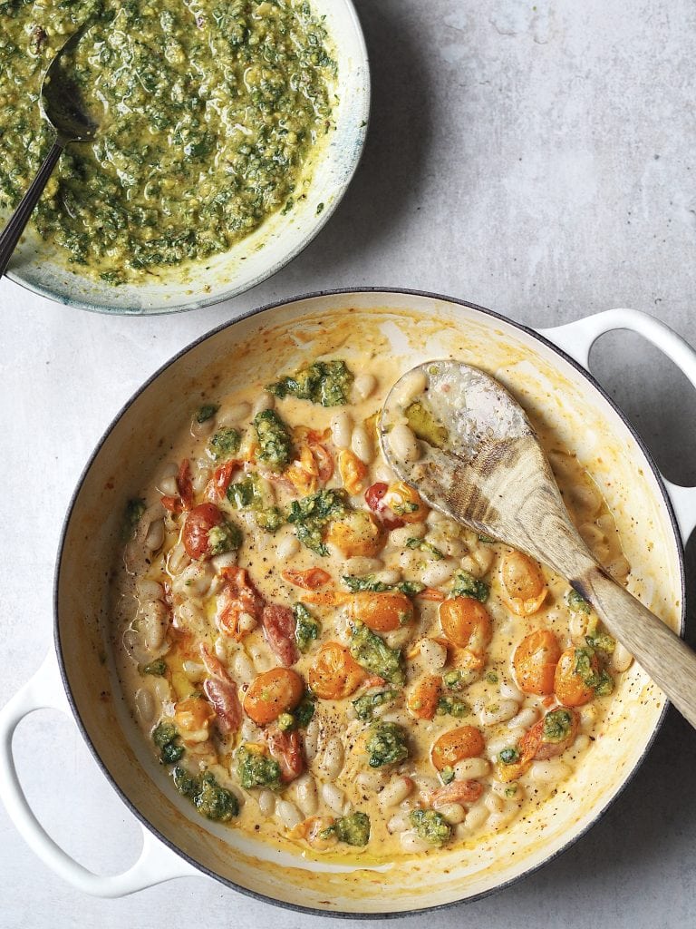 A pan of creamy white beans cooked with cherry tomatoes drizzled with pesto, in a bowl next to the pan.