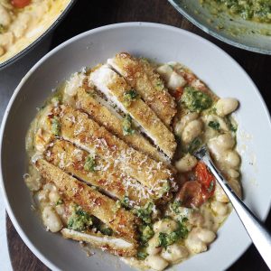 Grey bowl of white beans and cherry tomatoes topped with a sliced crispy chicken cutlet.