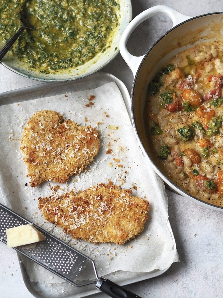 Tray of cooked crispy chicken cutlets topped with grated parmesan next to dishes of creamy tomato beans and homemade basil pesto.