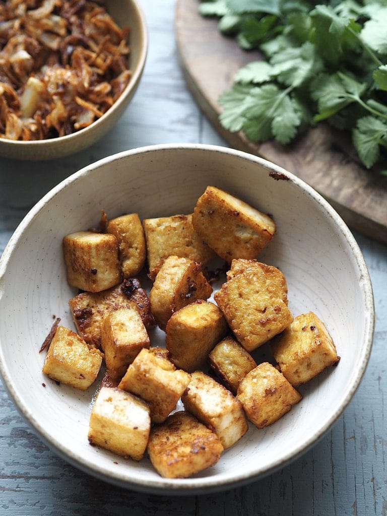 A stone bowl of crispy tofu with a bowl of crispy shallots and a bunch of coriander in the background.