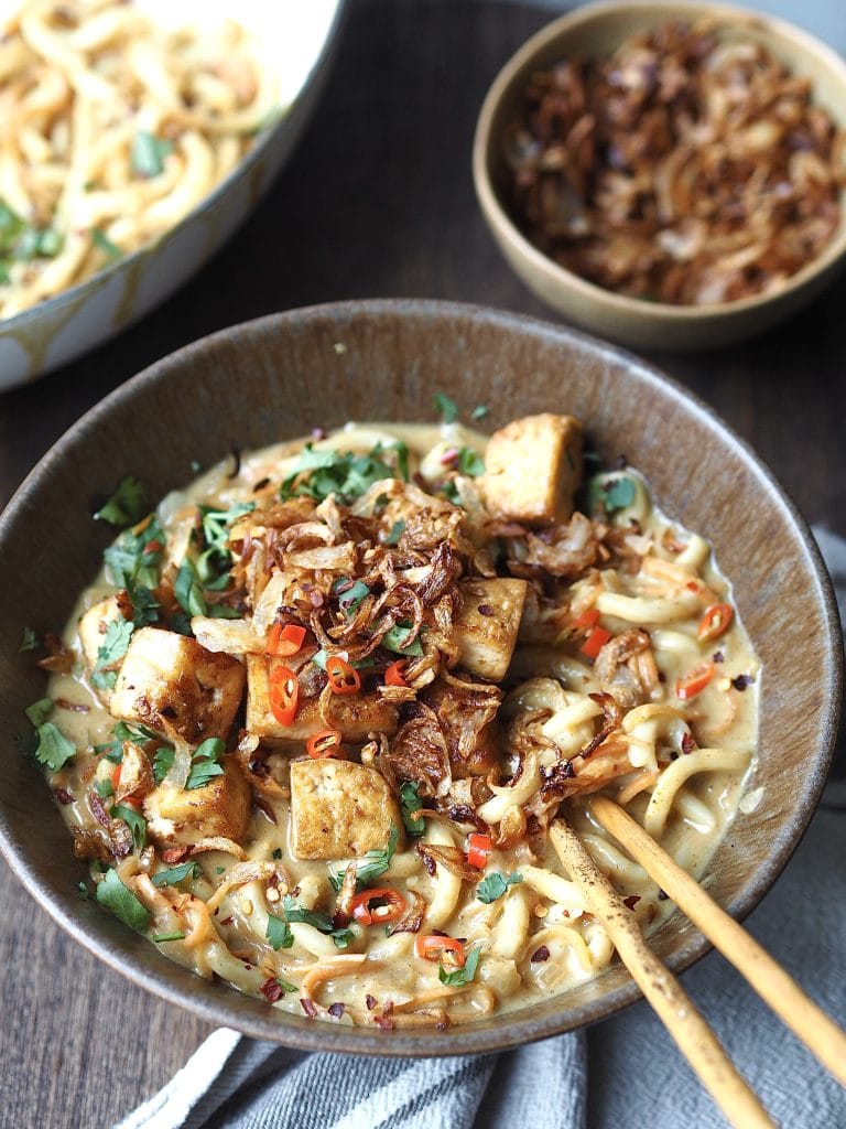 A close up of a bowl of curry udon noodles topped with crispy tofu cubes, fried shallots and sliced chilli.