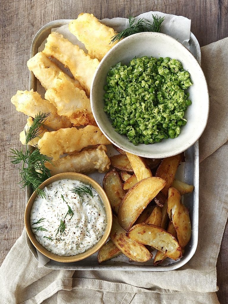 A metal tray of tempura fish and chips with bowls of smashed peas and homemade tartare sauce.
