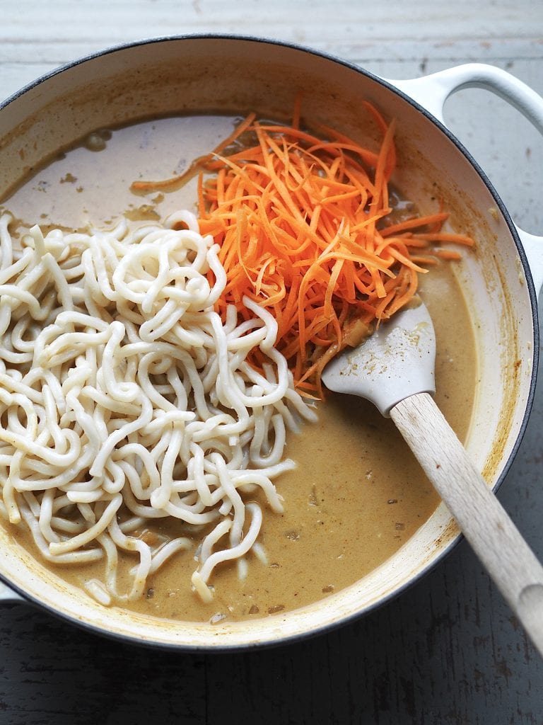 Shredded carrot and udon noodles being stirred into a creamy curry soup.