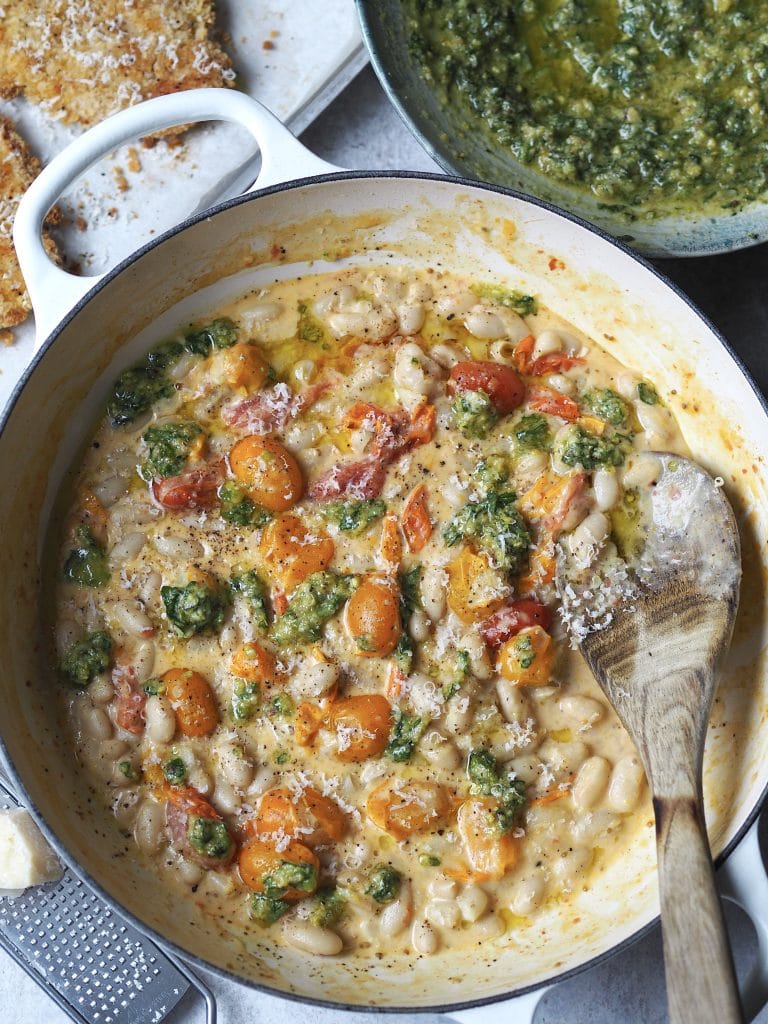 Close up of a white shallow casserole with creamy beans and cherry tomatoes drizzled with pesto.