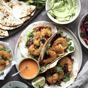 Plate of panko prawn tacos on soft corn tortillas surrounded with bowls of fixings.