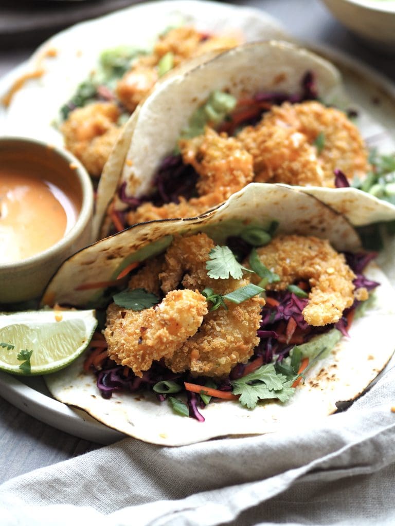 Close up of panko prawn tacos with lime wedges and a bowl of sriracha sauce on the side.