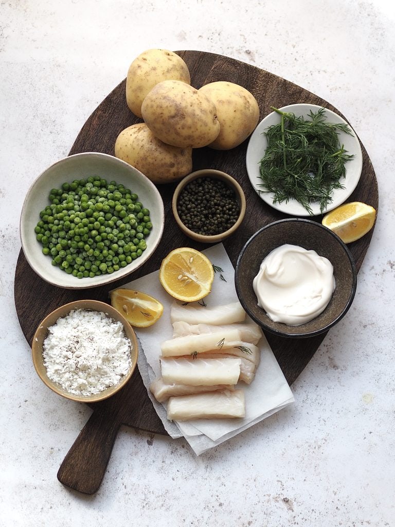 Wooden board with all the ingredients to make fish goujons with smashed minty peas, potato wedges and tartare sauce.