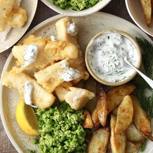 Fish goujons on a plate with potato wedges, lemon and peas drizzled with homemade tartare sauce.