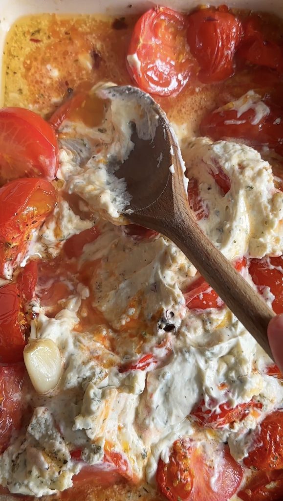 a wooden spoon mashing roasted tomato with boursin