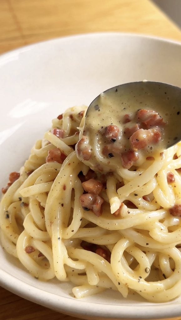 spooning creamy sauce over the udon carbonara