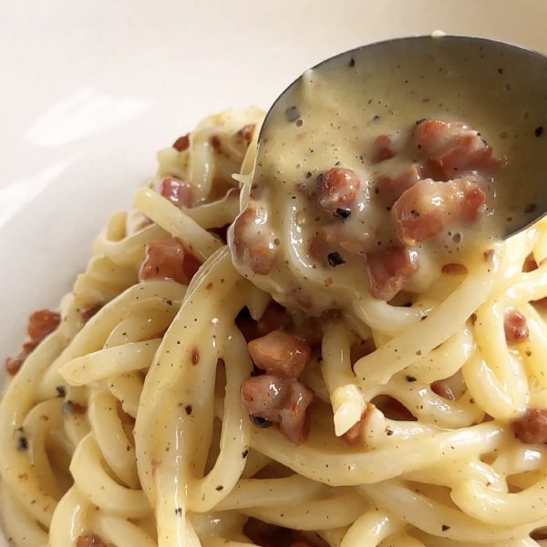 spooning creamy sauce over the udon carbonara