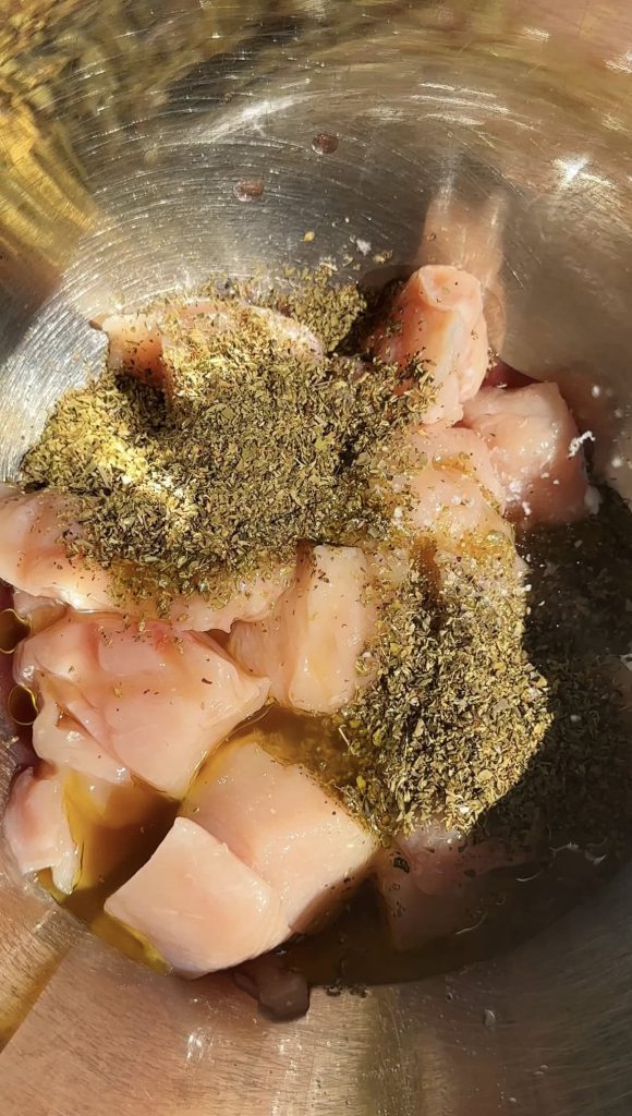 adding dried herbs and marinade to cubed chicken breast in metal bowl