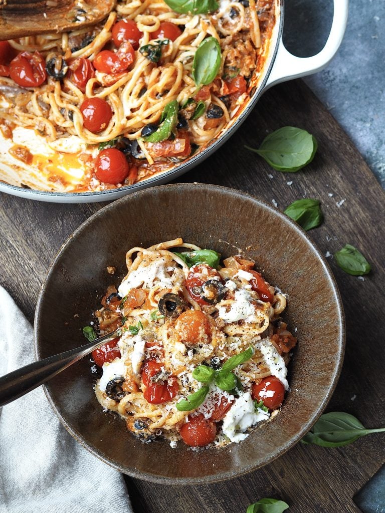 Bowl of pasta with burrata, cherry tomatoes and black olives topped with fresh basil next to the casserole dish filled with pasta.