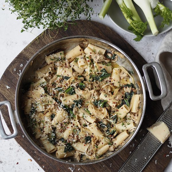 Cooking pot filled with creamy sausage and kale pasta on a wooden board.