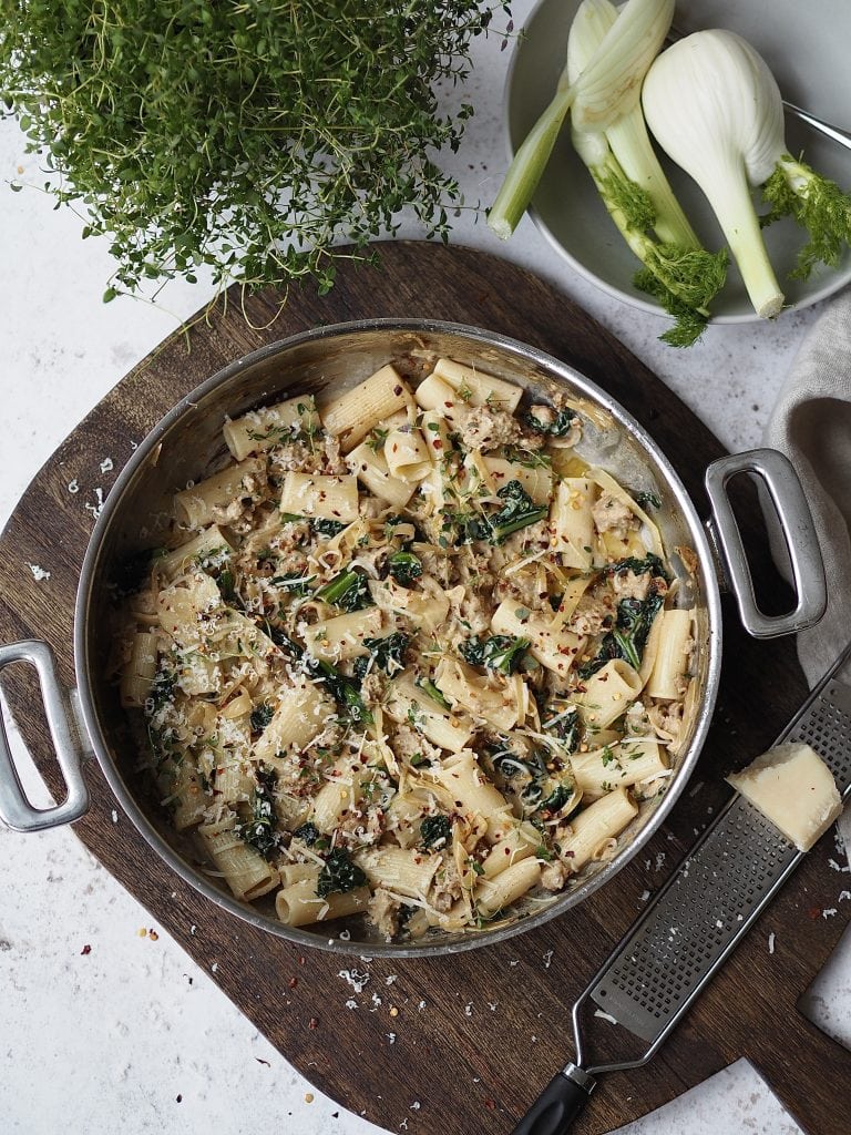 Cooking pot filled with creamy sausage and kale pasta on a wooden board.