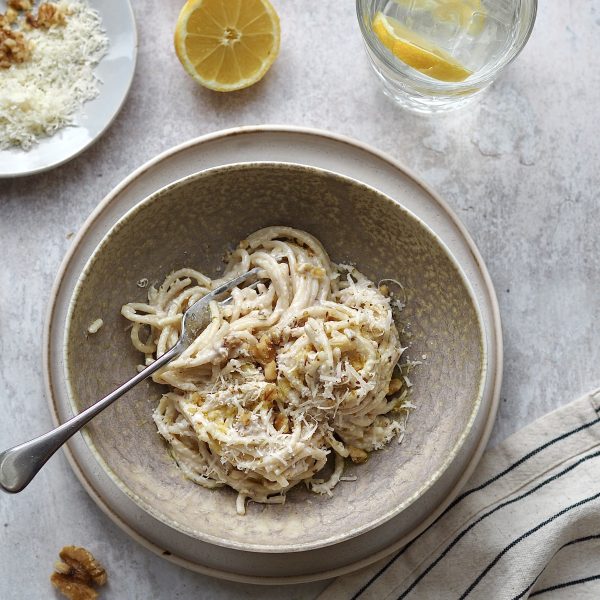 Grey bowl of spaghetti in a white sauce surrounded by a glass of water and lemon halves.