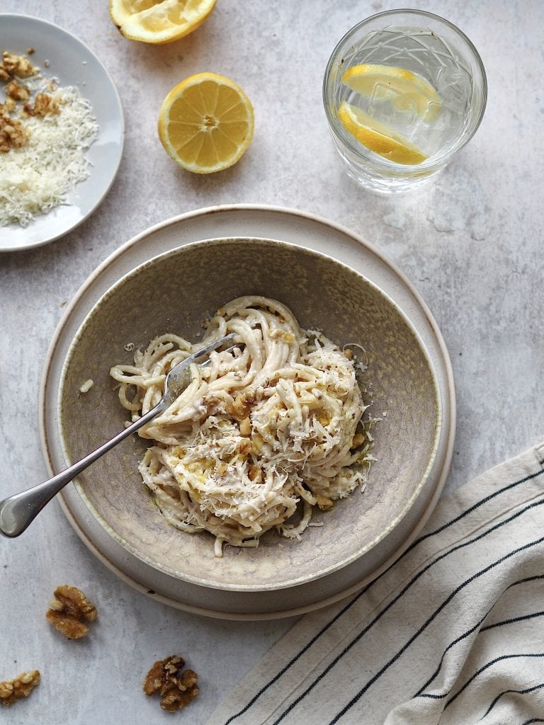 Grey bowl of spaghetti in a white sauce surrounded by a glass of water and lemon halves.