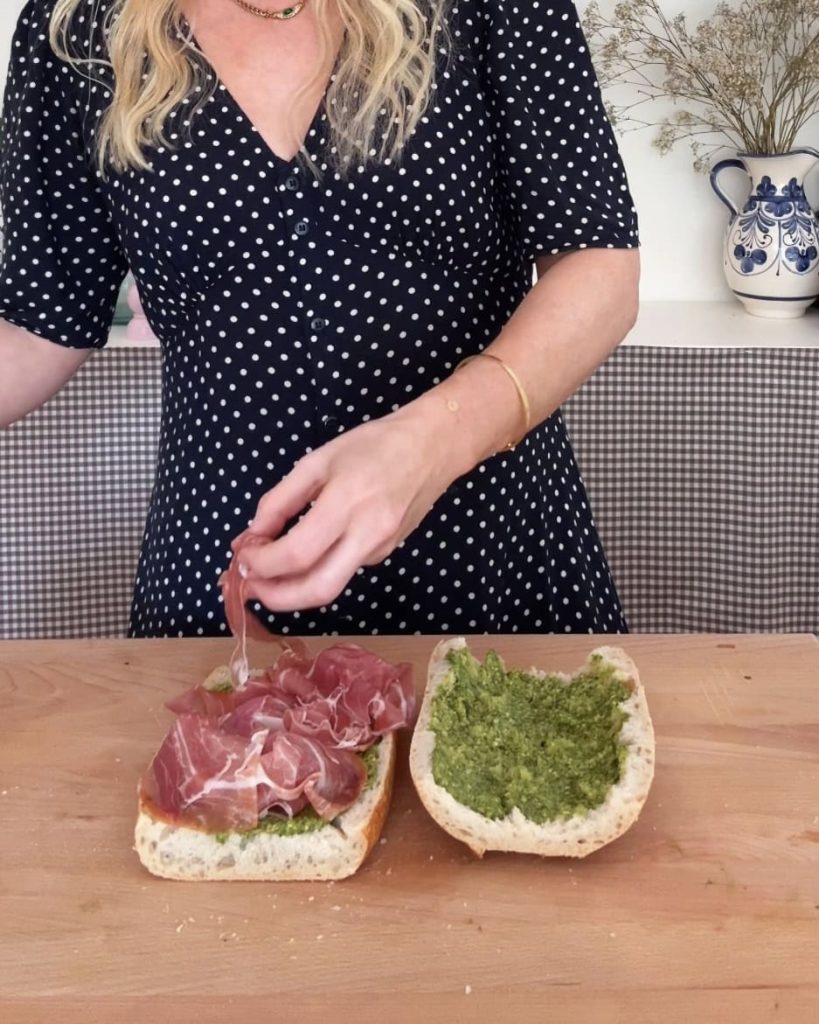 woman adding prosciutto to bread with pesto spread