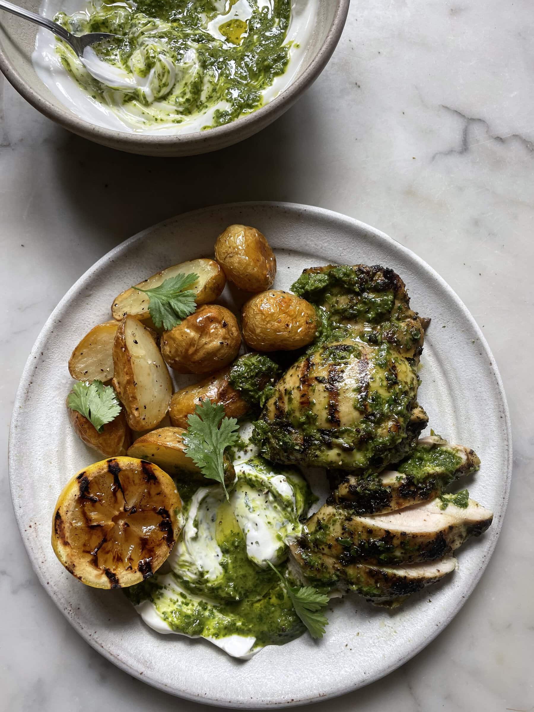 Zingy Grilled Chimichurri Chicken with Roasted Potatoes on a grey plate with a bowl of chimichurri yoghurt behind