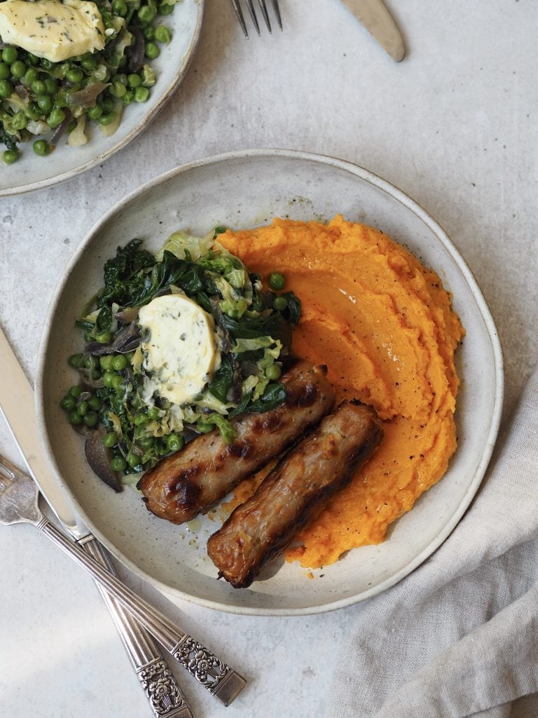 Grey plate of sweet potato mash, roasted sausages, wilted greens and garlic butter.