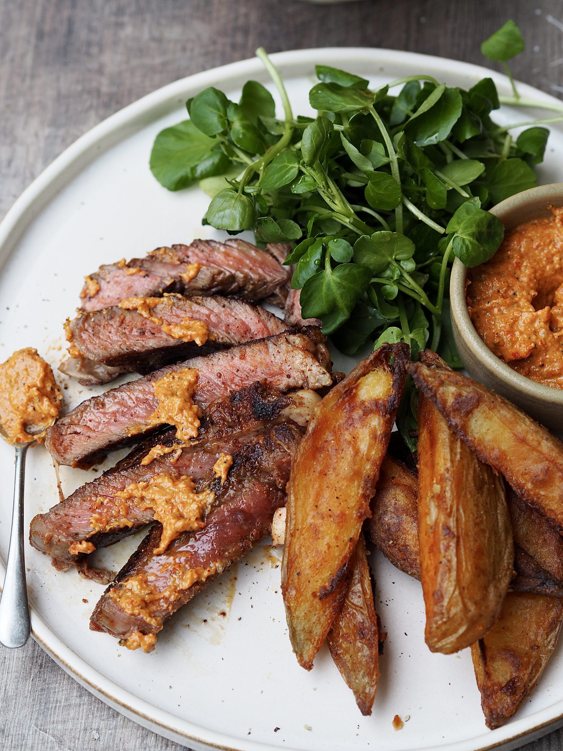 White plate of steak with romesco sauce, watercress and potato wedges.