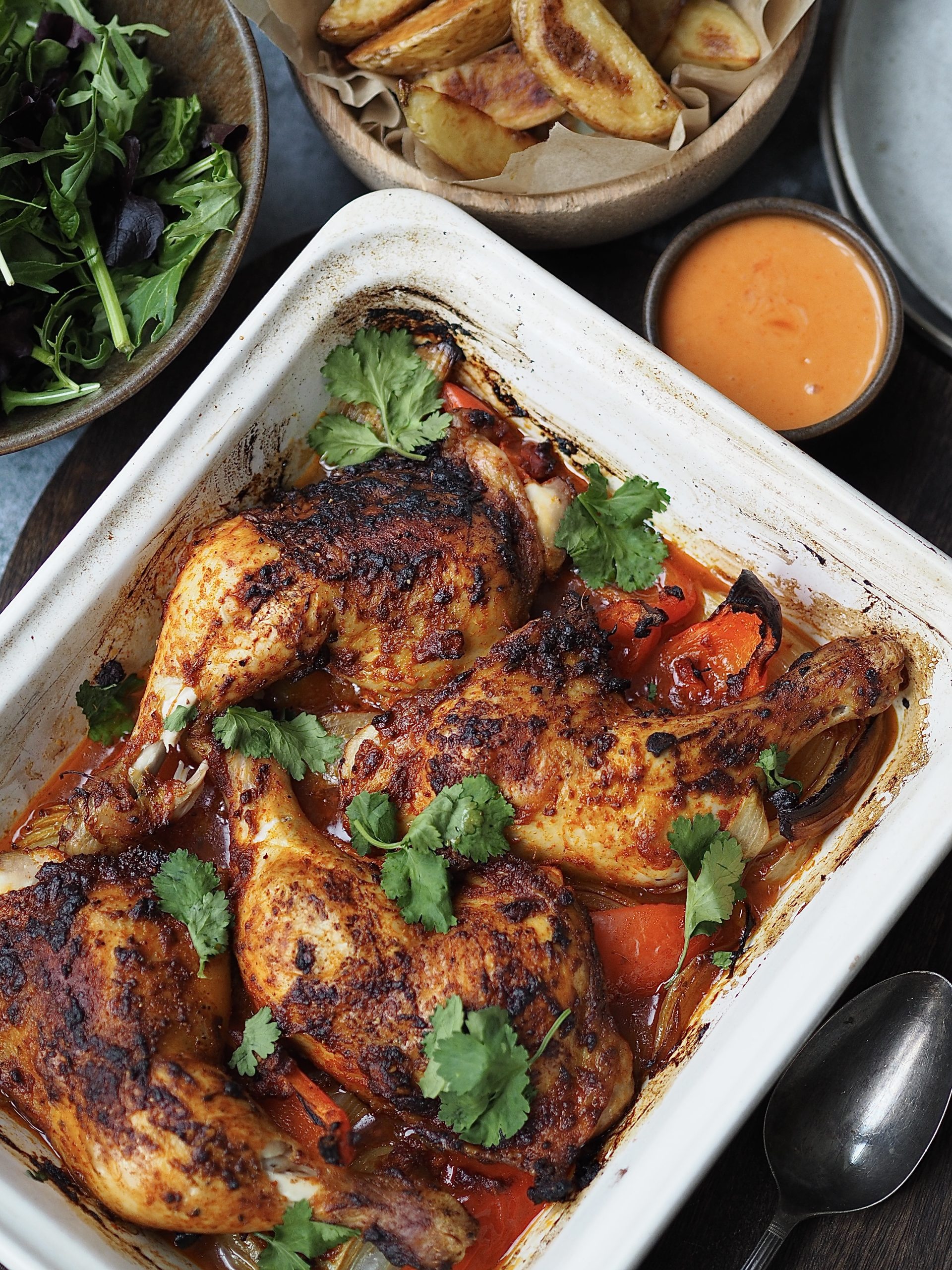 Close up of a white roasting dish with peri peri chicken legs garnished with fresh herbs, with bowls of potato wedges, sriracha mayo and green salad leaves off to one side.