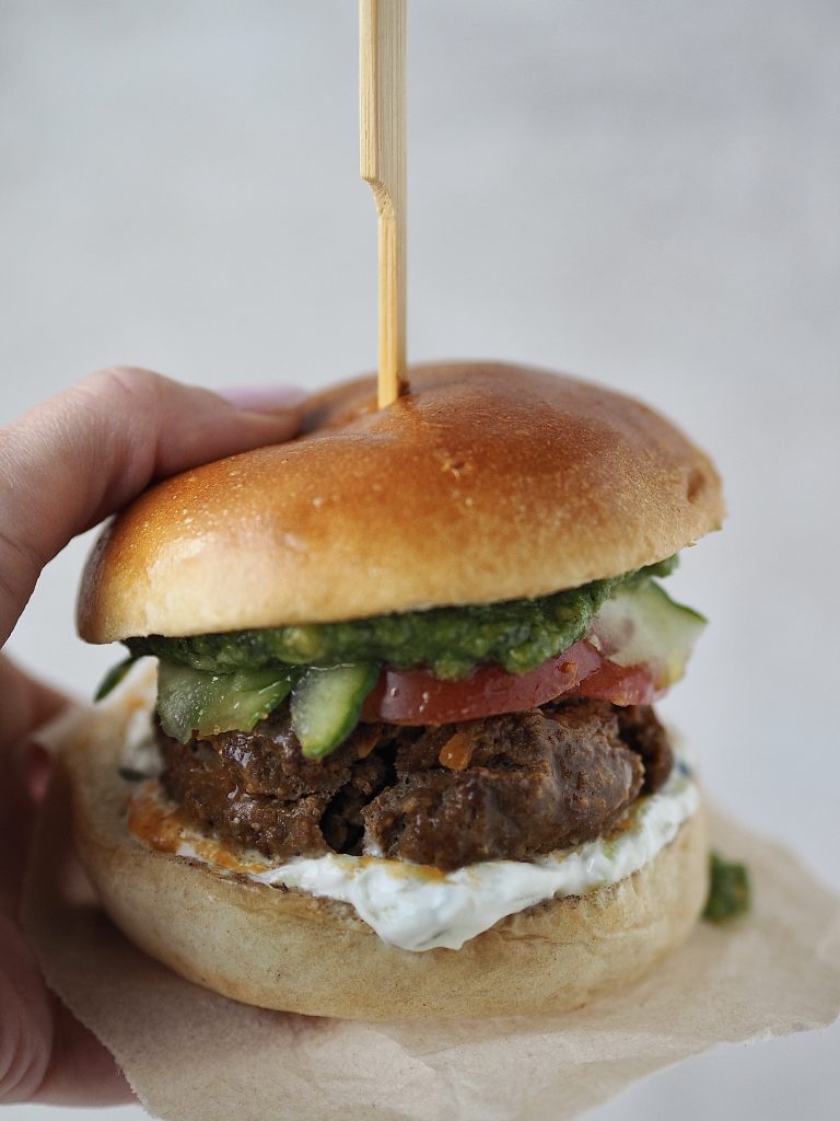 Hand holding a lamb burger in a brioche bun with salad, tzatziki and pesto.