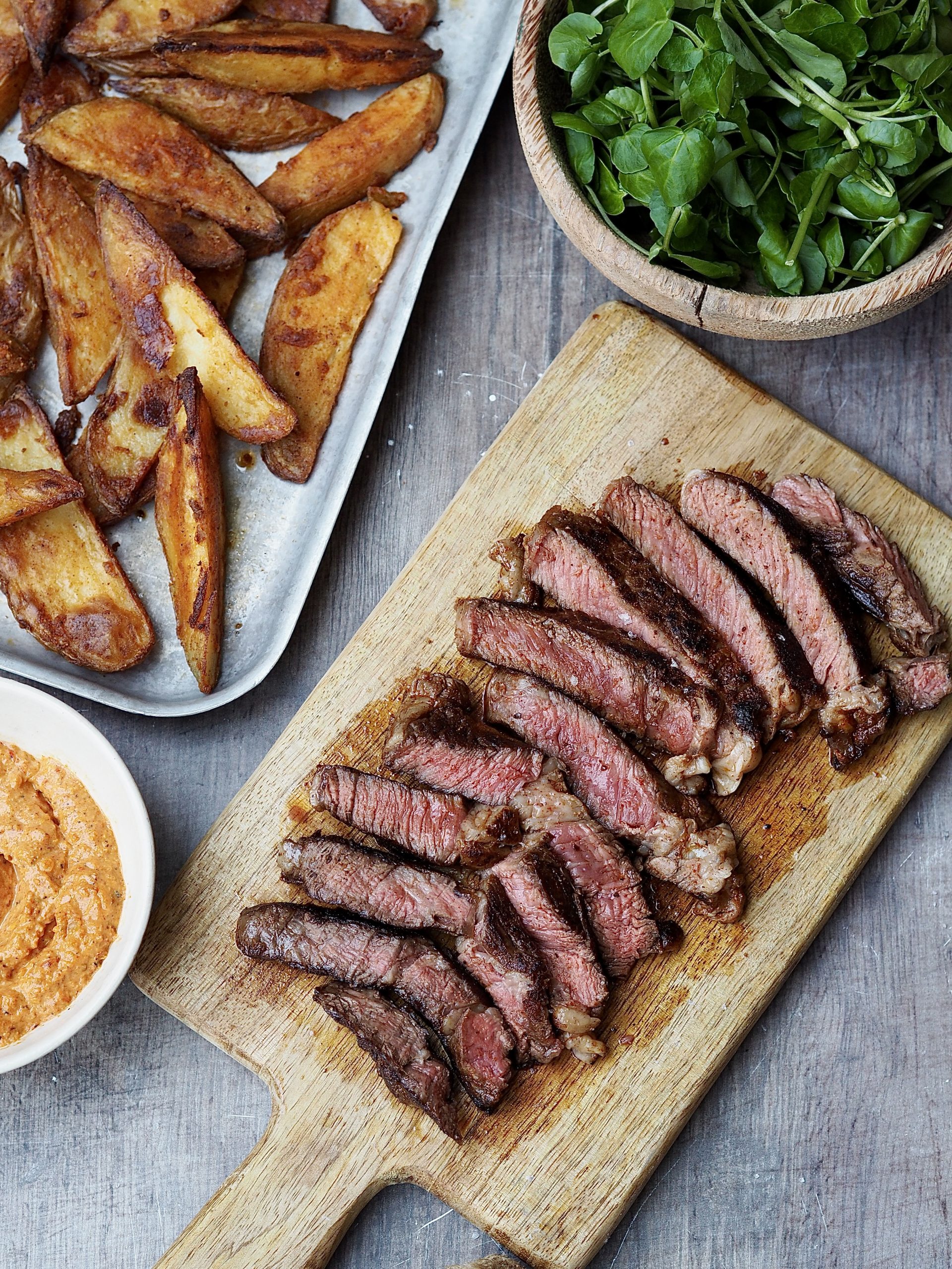 Sliced steak on a cutting board with a tray of potato wedges and bowls of romesco and watercress on the side.