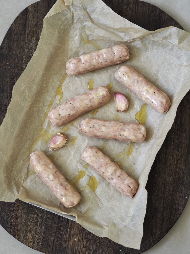 Sausages and whole garlic cloves drizzled with oil on a baking parchment lined baking tray.