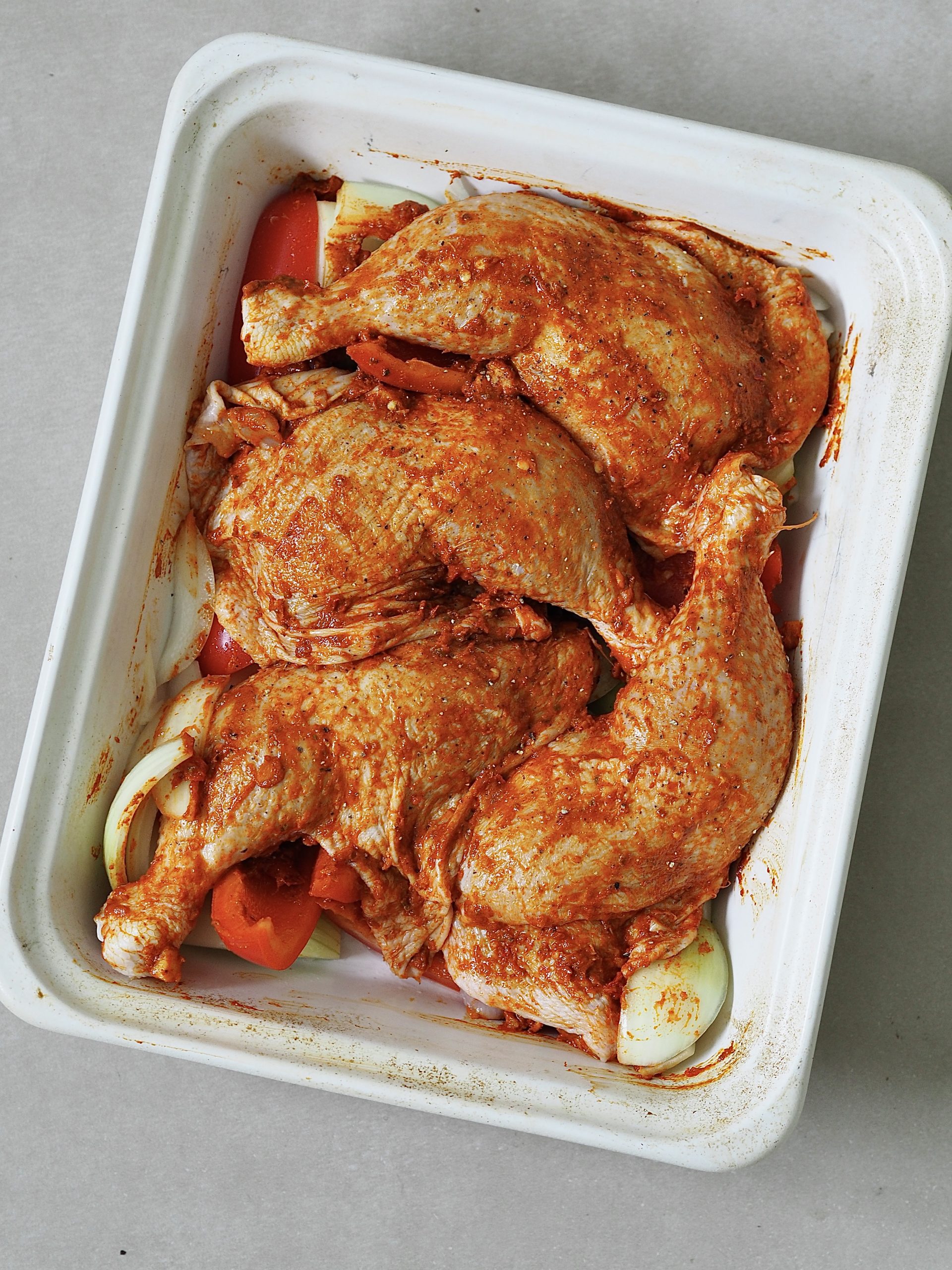 Peri peri marinated raw chicken legs in a white roasting tray on a bed of red peppers and onions.