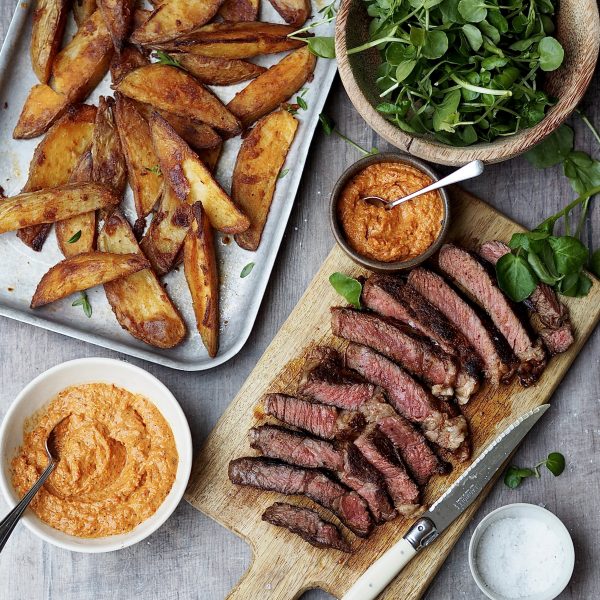 Trays of crispy potato wedges and sliced steak next to bowls of watercress and romesco sauce.