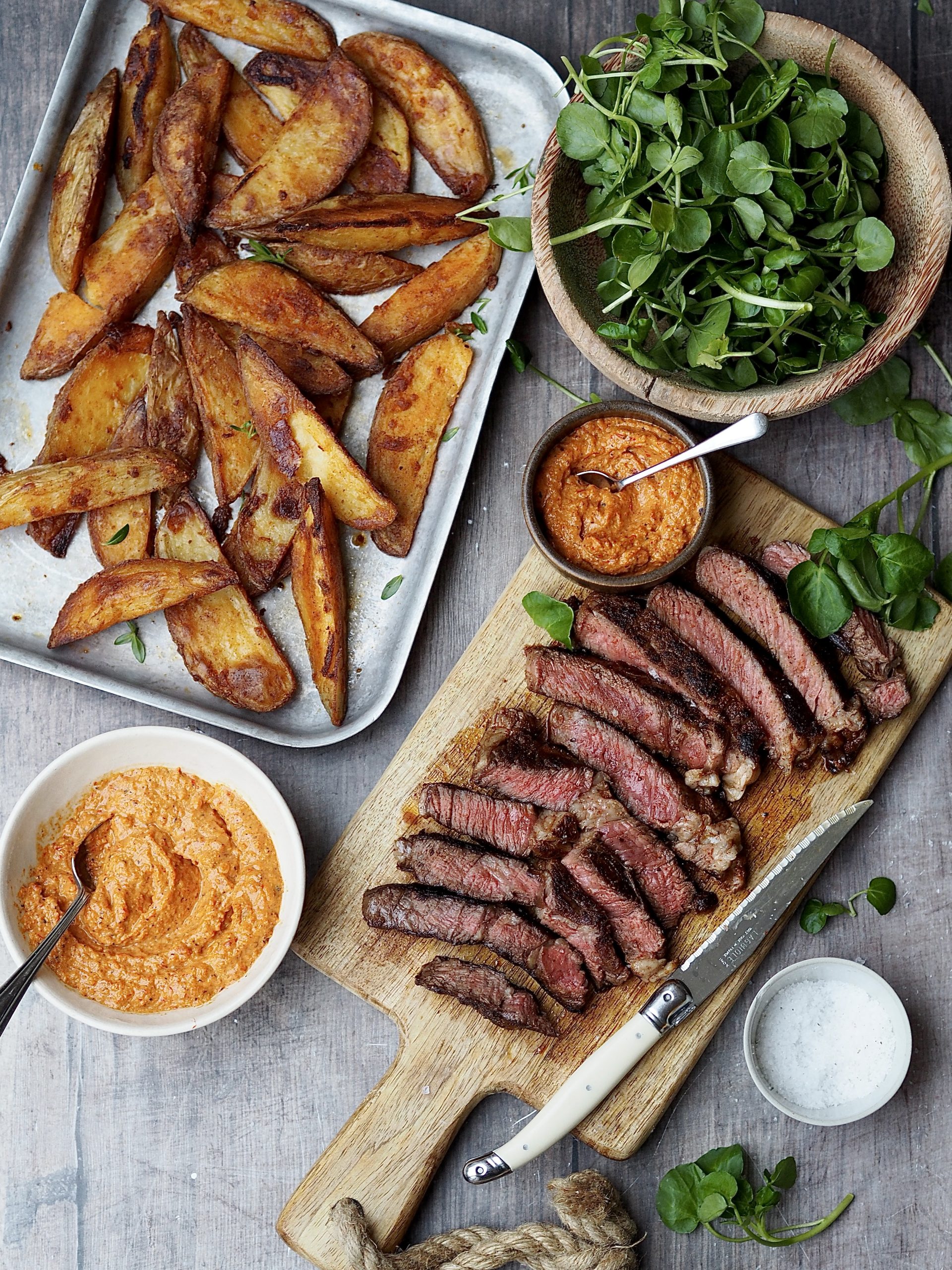 Trays of crispy potato wedges and sliced steak next to bowls of watercress and romesco sauce.