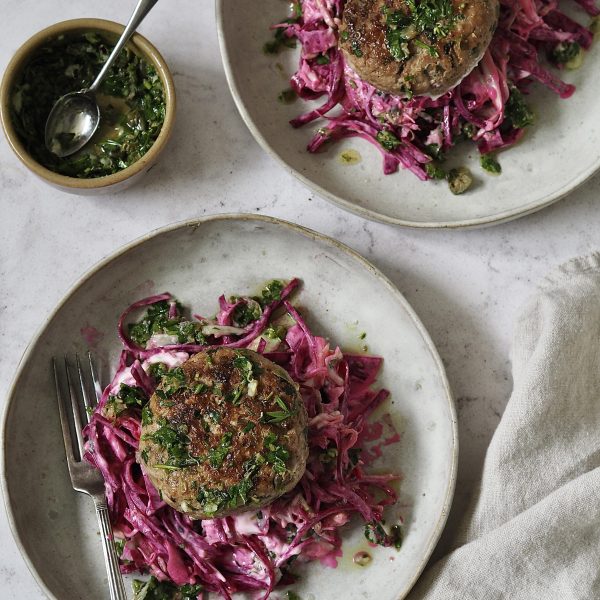 Chicken burgers on piles of pink yoghurt and beetroot slaw on grey stone plates next to a small bowl of salsa verde that's been spooned over the top.