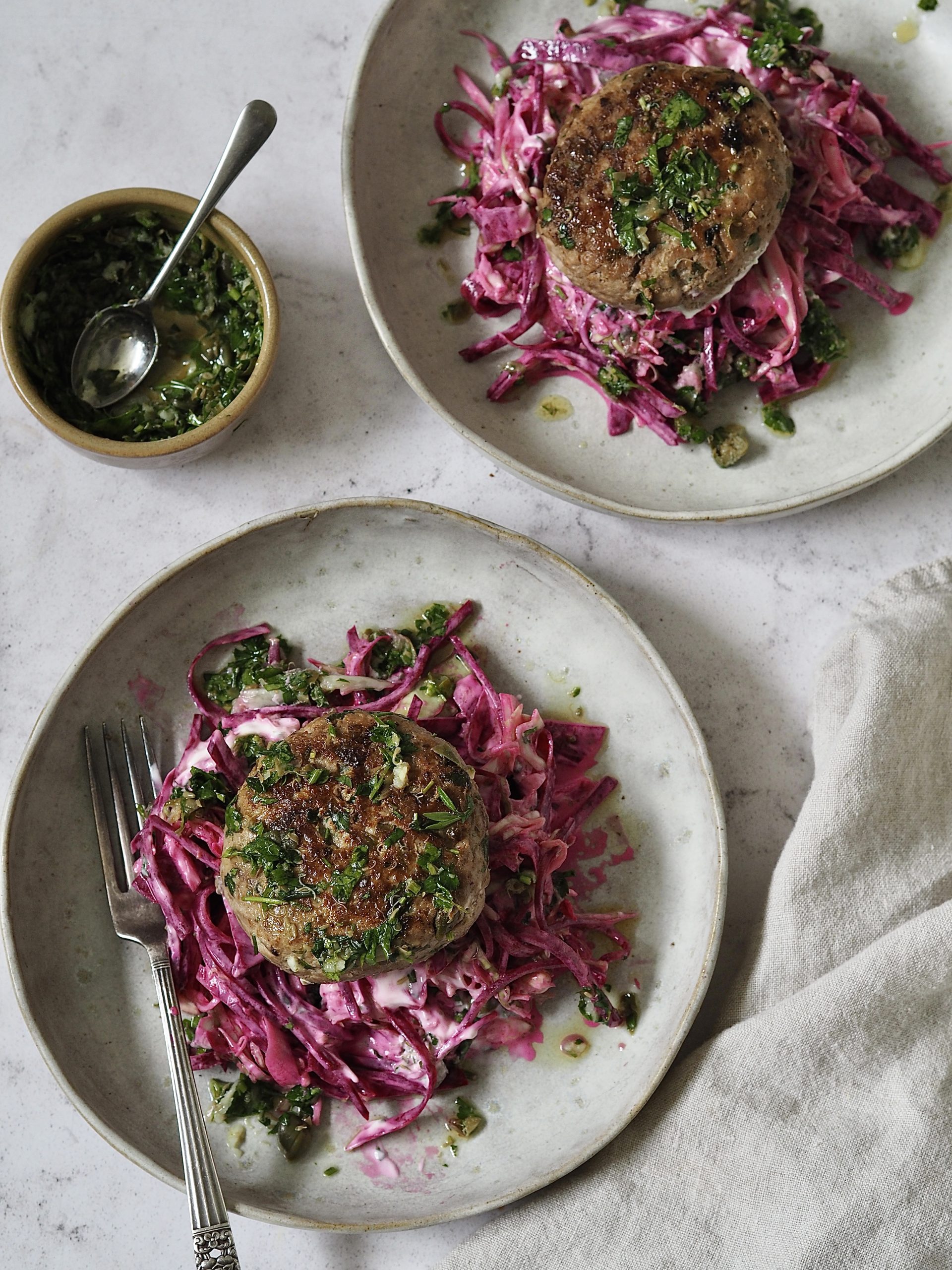 Chicken burgers on piles of pink yoghurt and beetroot slaw on grey stone plates next to a small bowl of salsa verde that's been spooned over the top.