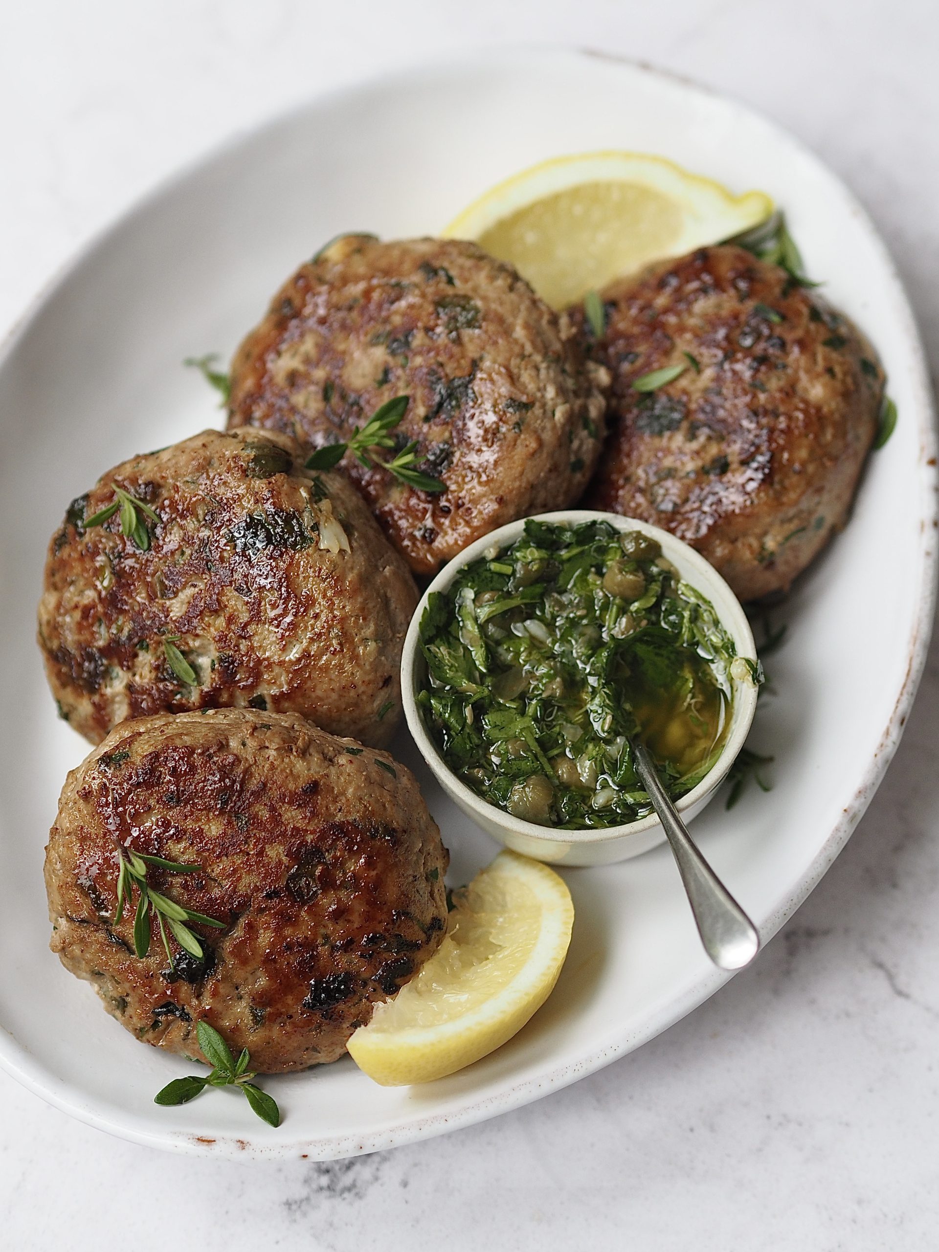 Chicken burgers on a white serving platter with lemon wedges, fresh herbs and a small white bowl of salsa verde.