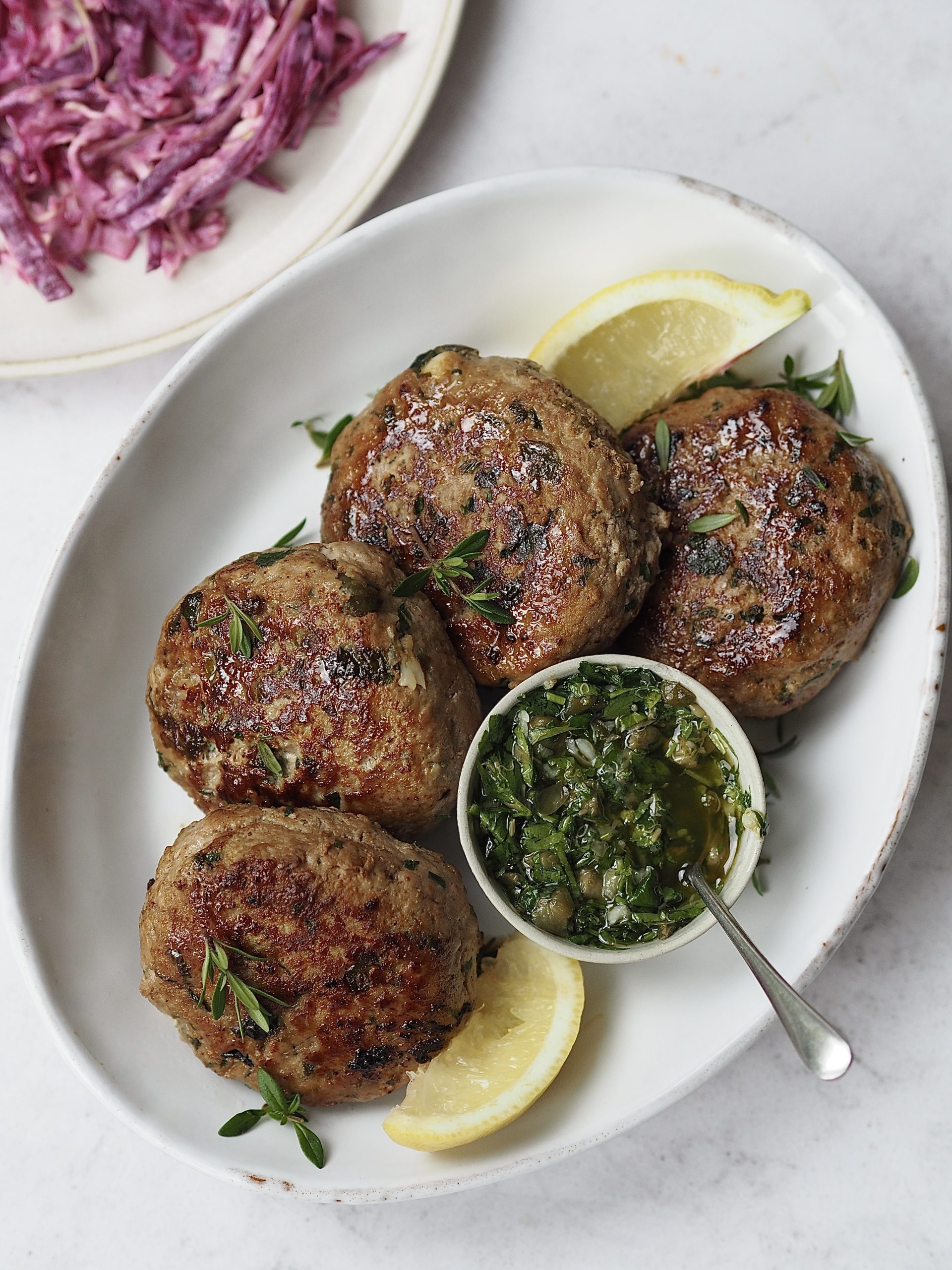 White plate of herby chicken burgers with lemon wedges, a small bowl of salsa verde and a plate of yogurt slaw on the side.