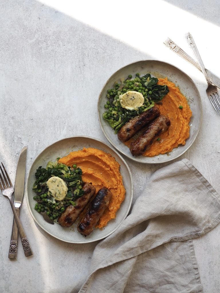 Plates of bangers, sweet potato mash and greens on a grey countertop.