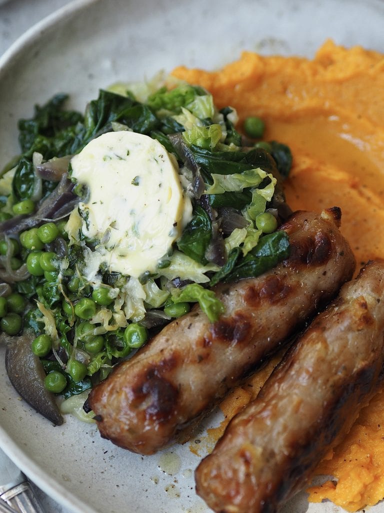 Close up of roasted sausages served with greens topped with garlic butter and some mashed sweet potato.