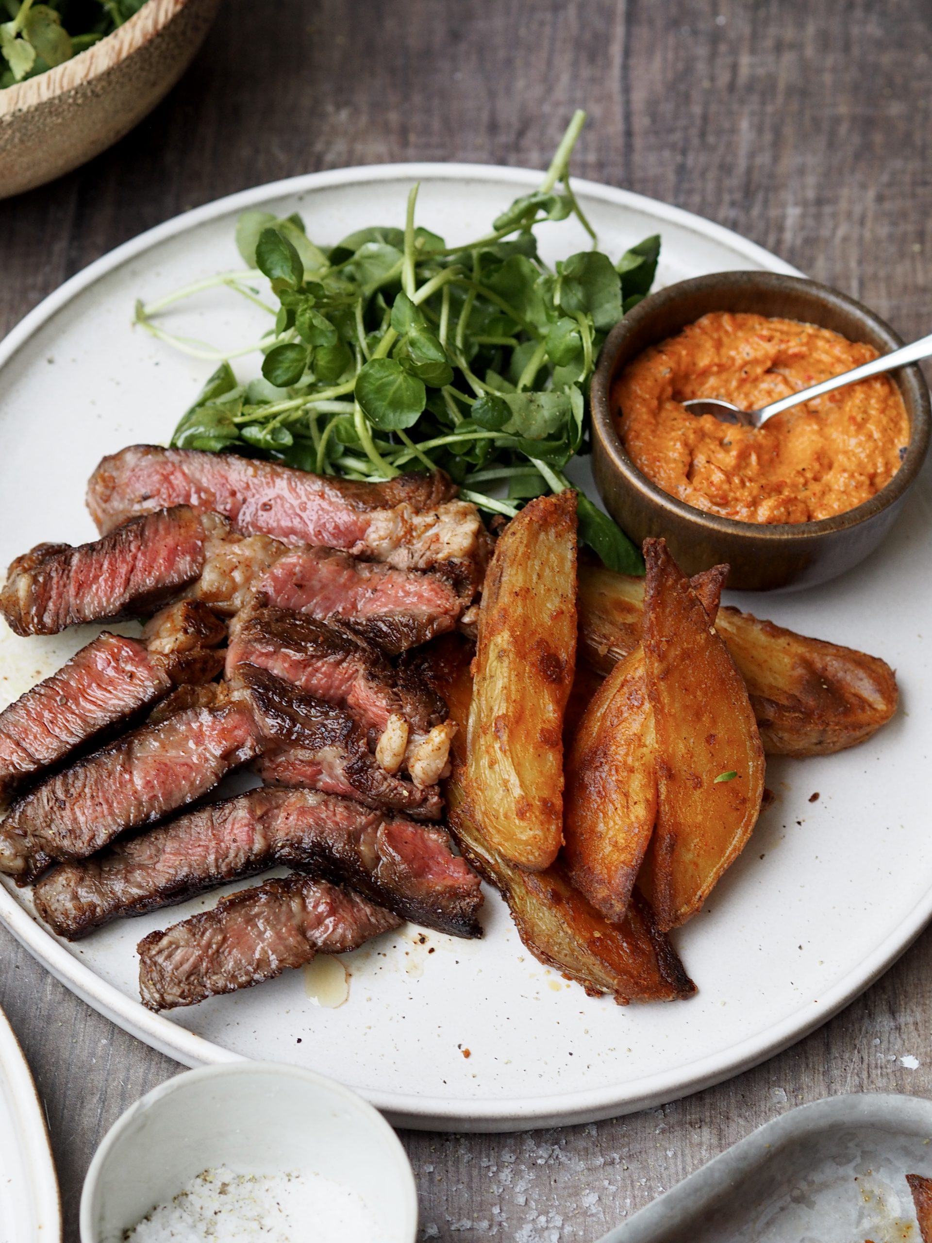 White plate of sliced steak, potato wedges and watercress with a little black bowl of romesco sauce. 